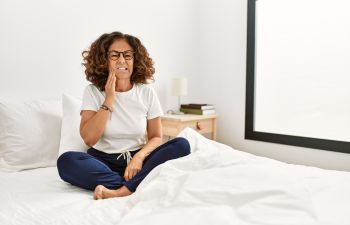 Middle-aged Hispanic woman suffering from bruxism sitting on her bed and touching her jaw .