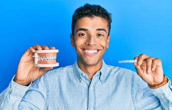 Young Afro-American man with a perfect smile holding traditional braces in one hand and a clear aligner in the other one.