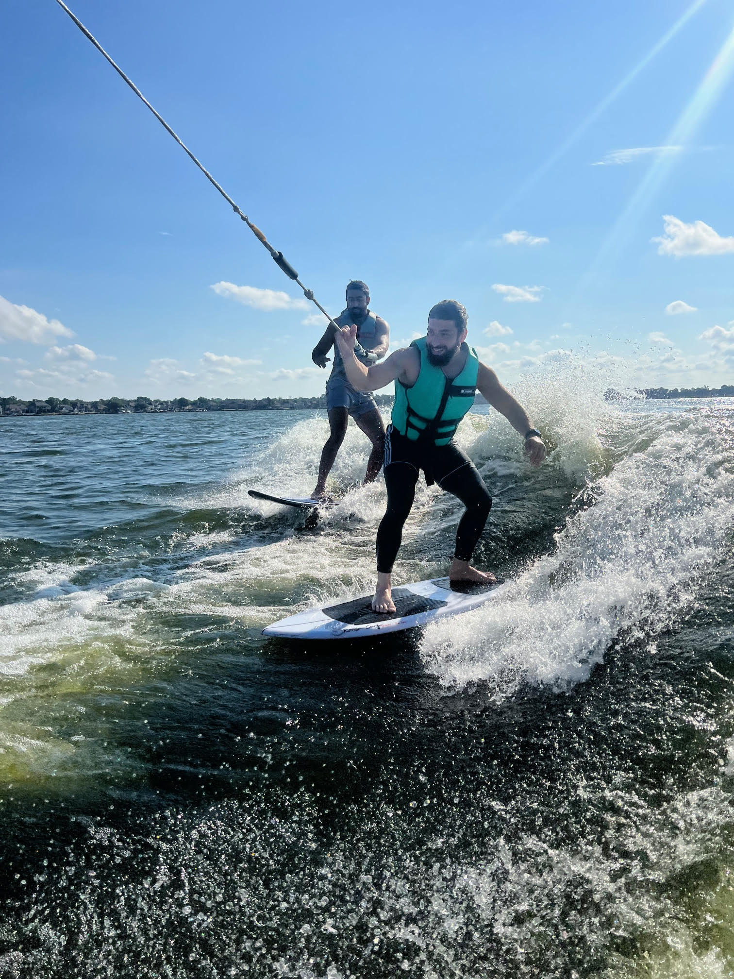 Two people ride surfboards on the water while being towed by a rope. Both wear life vests, with waves splashing around them under a sunny sky.