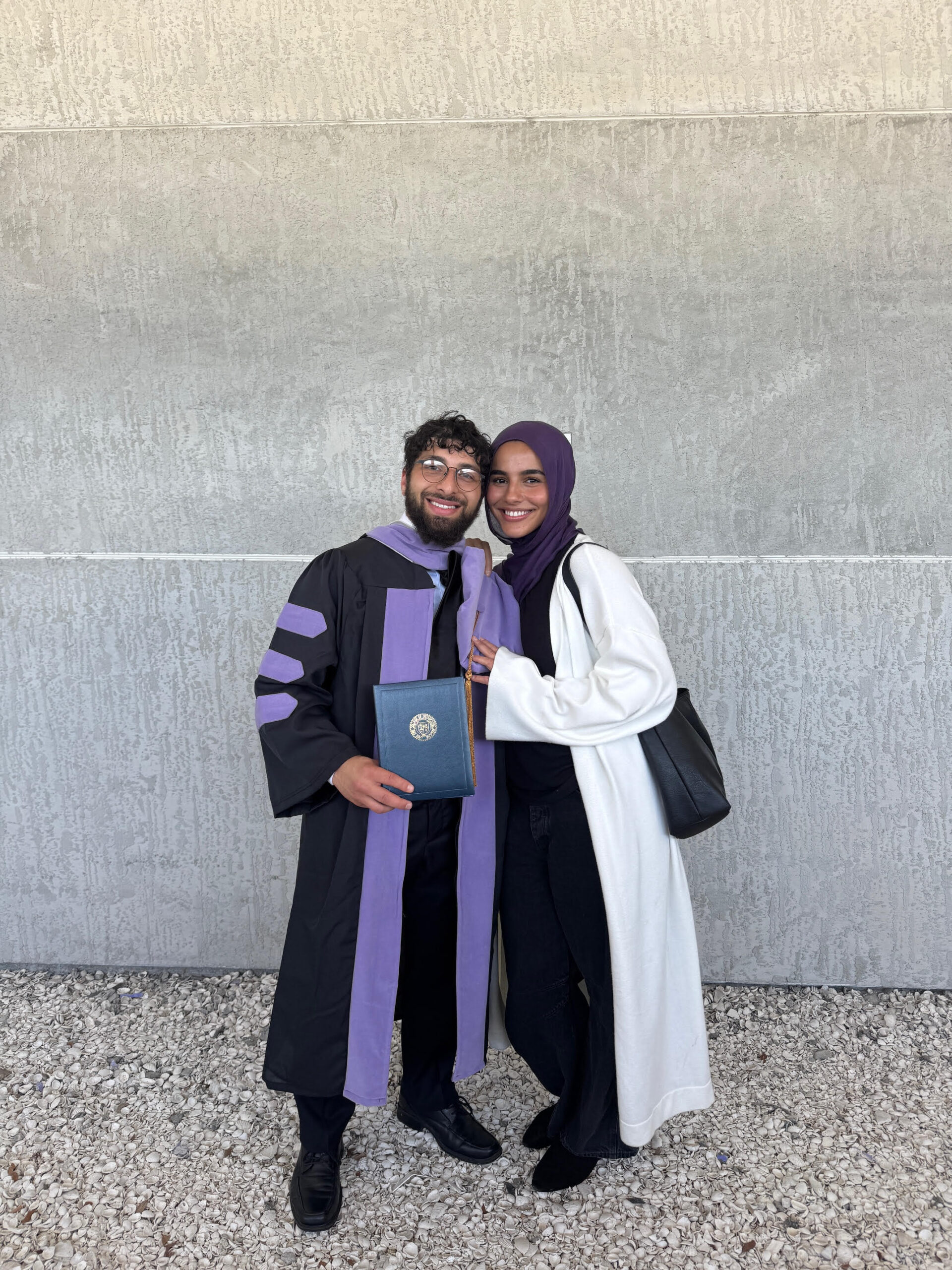 A graduate in a black and purple gown stands beside a woman wearing a white coat and headscarf. The graduate holds a diploma folder, and both smile in front of a light-colored wall with gravel beneath their feet.