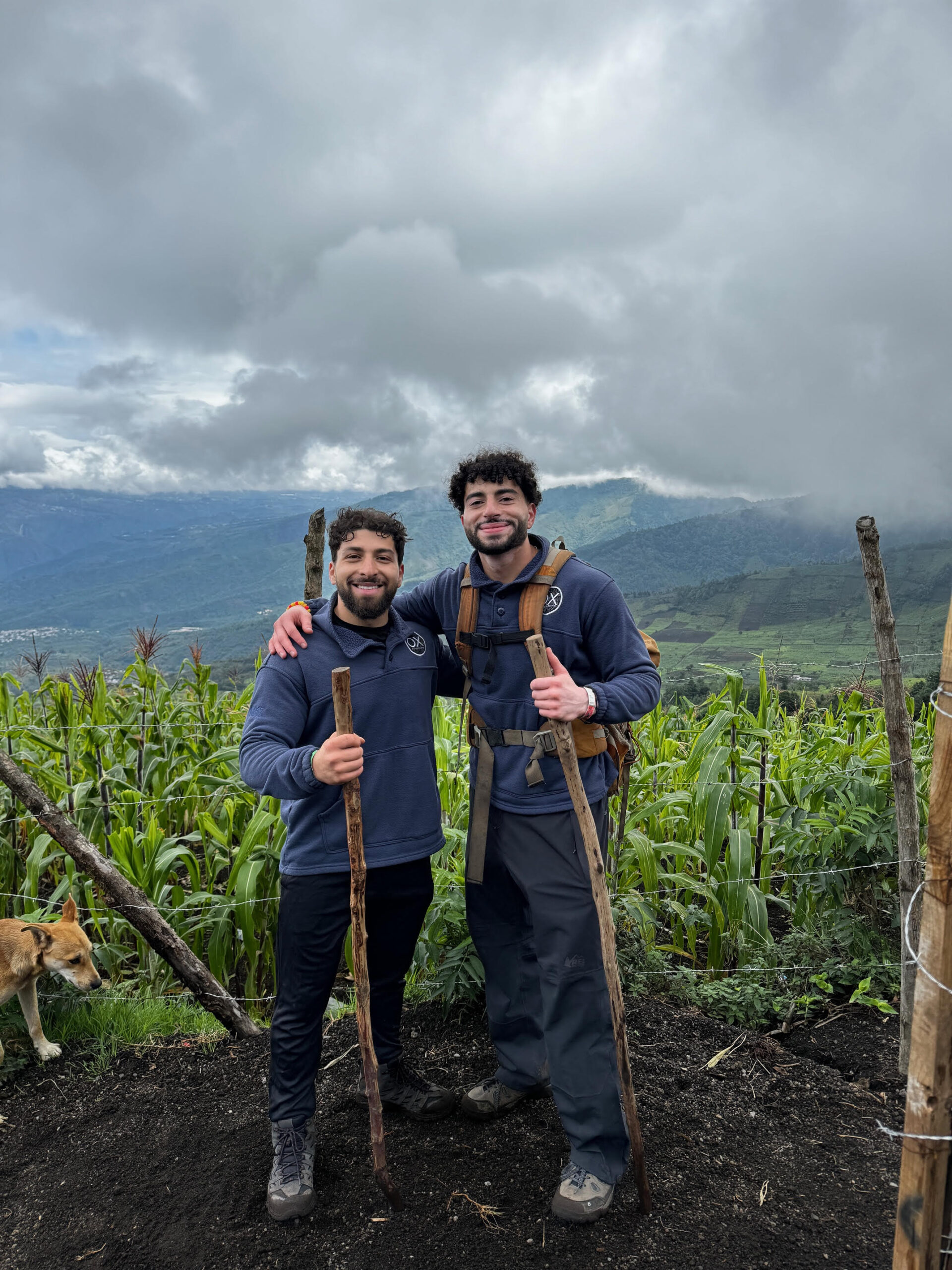 Two men pose next to each other on a mountain trail, holding wooden walking sticks and wearing outdoor clothing. Lush green crops and distant hills are visible in the background, with a dog nearby.