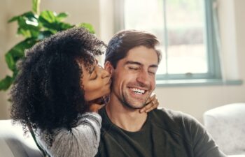 Happy man, woman and kiss on cheek on sofa for love, affection and proud for engagement. Interracial couple, hug and commitment in relationship with good news, trust and support at home with care